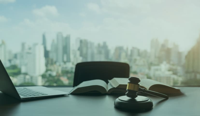 Gavel on desk with open books and laptop, overlooking a blurred city skyline through large window.